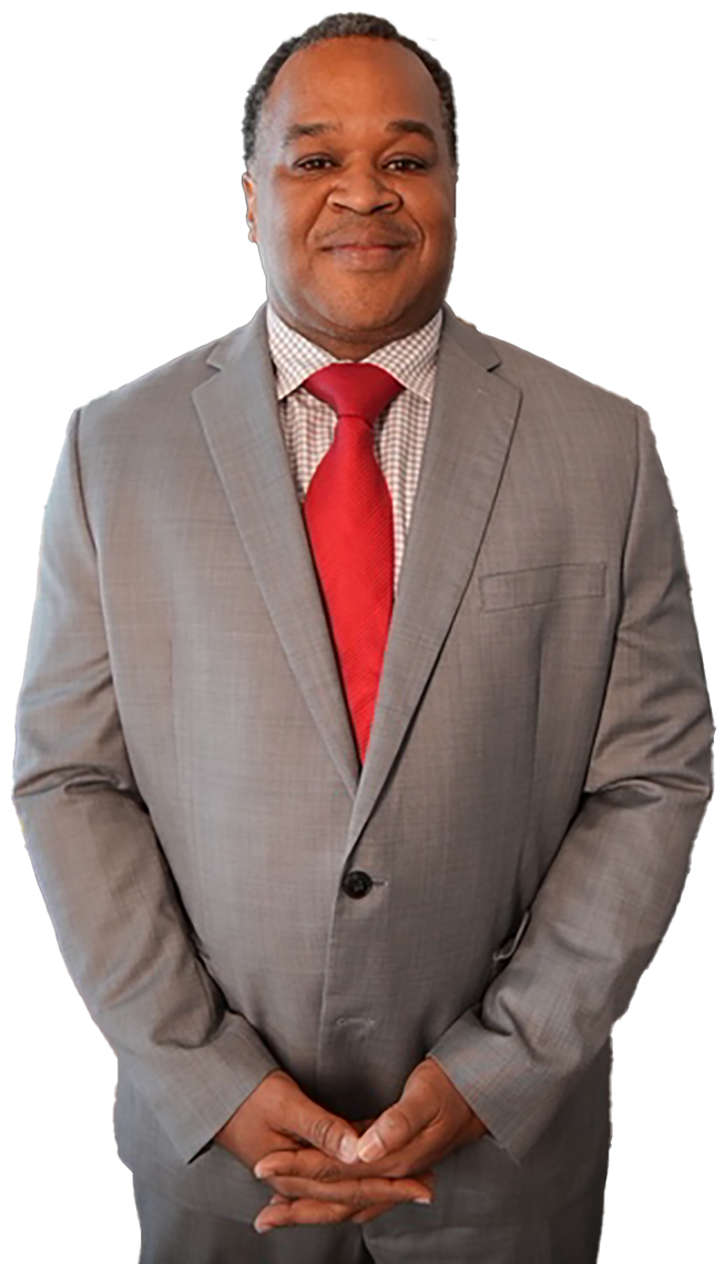 A man in a grey suit, patterned dress shirt, and red tie stands facing forward with his hands clasped against white.