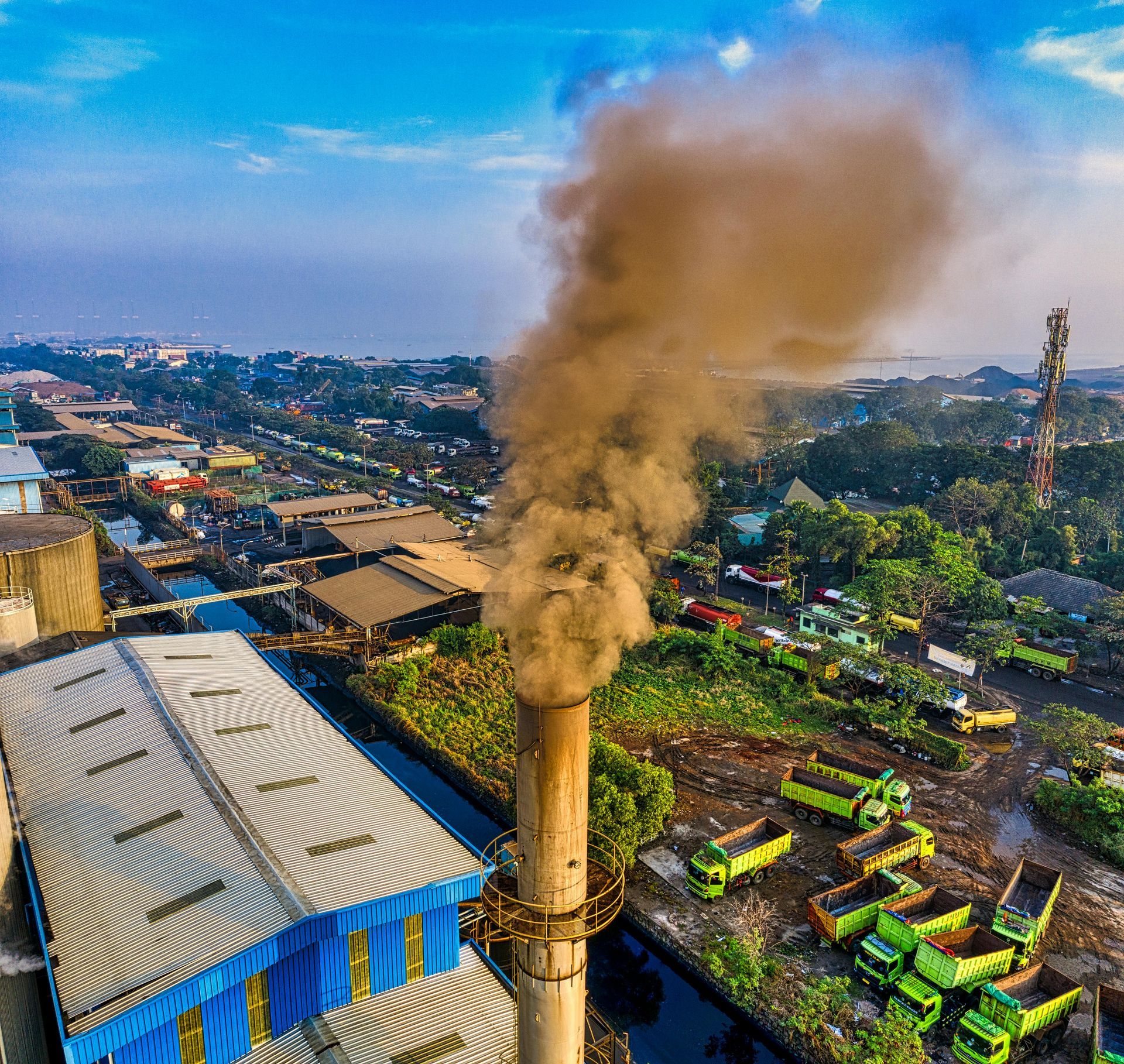 An industrial chimney emits a dense plume of dark smoke over a facility with a row of parked green dump trucks.