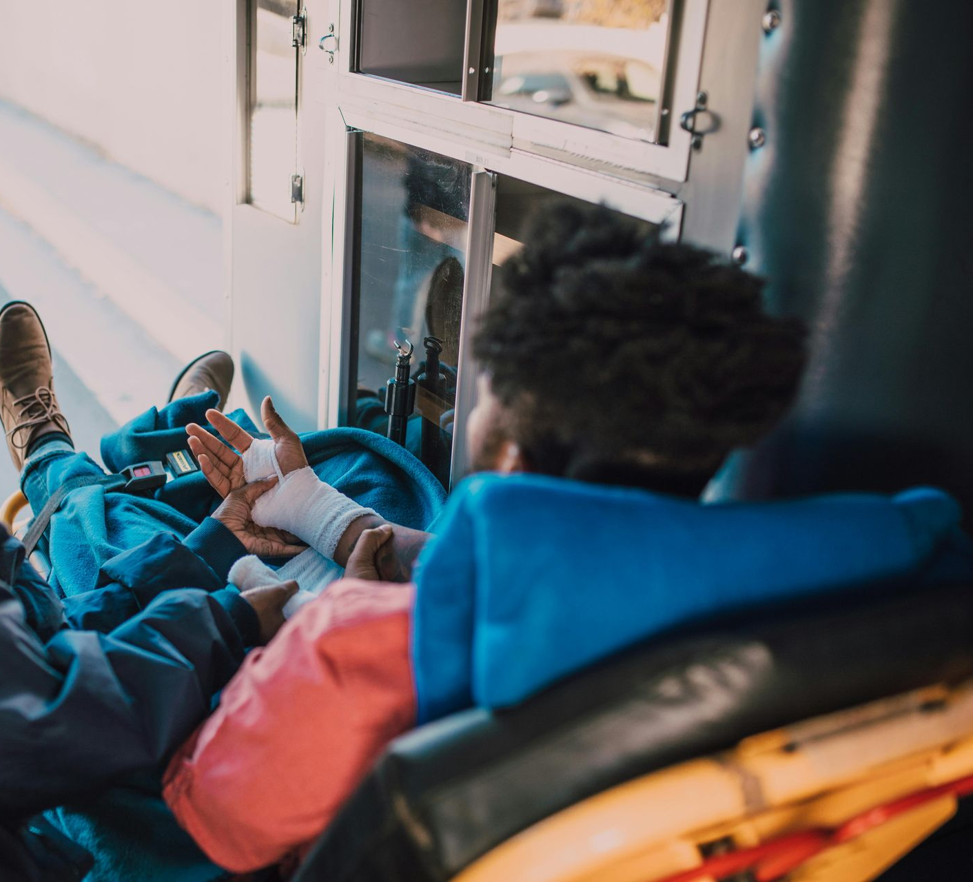 A person with a bandaged hand resting on their lap while sitting in a transport vehicle, wearing a pink and blue jacket.