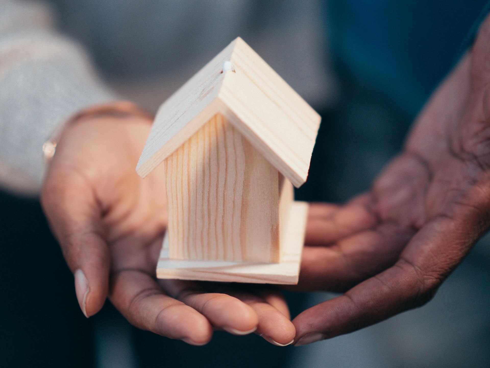 Two people cupping a small, light-colored wooden house model between their hands.