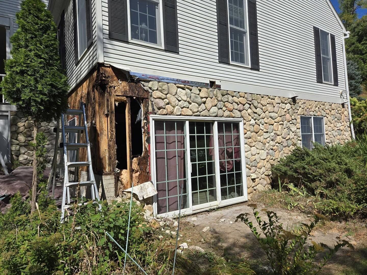Stone-and-siding house with damaged corner porch, white sliding doors, and overgrown garden.