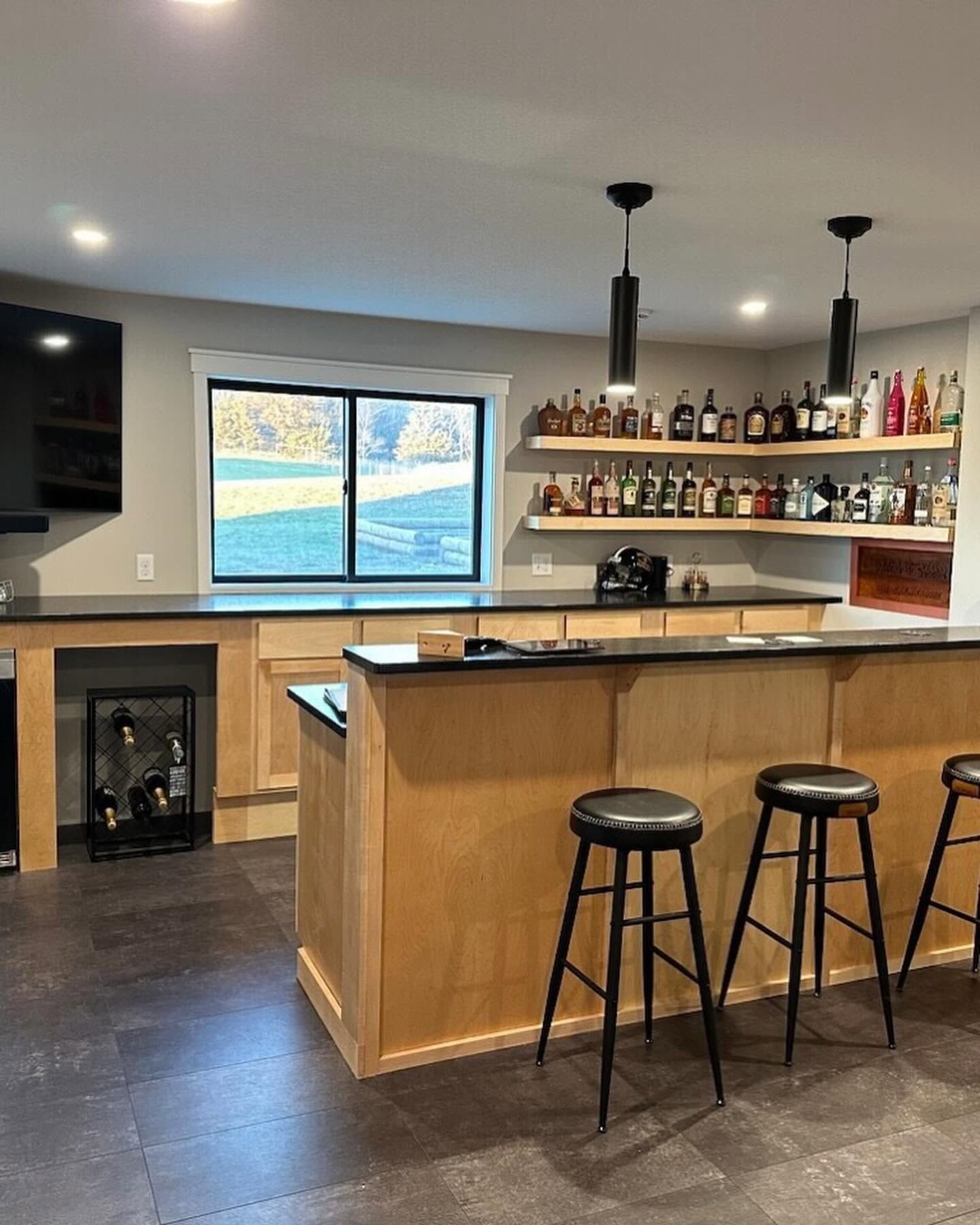 Modern kitchen bar with wooden island, three stools, shelves of bottles, and a window above the sink.