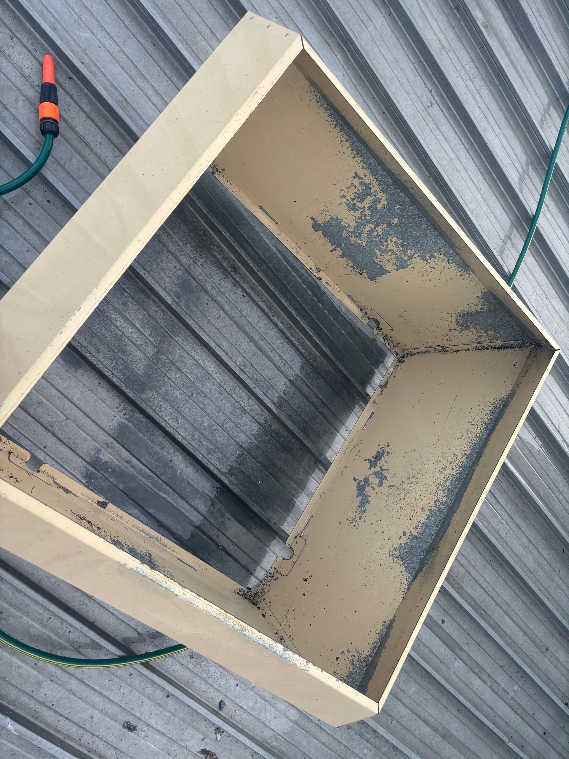 Square wooden box on a corrugated metal surface. Dark stains line the inside. A hose and nozzle are visible. -  DM & MM Services, Exhaust Cleaning, Townsville, QLD.