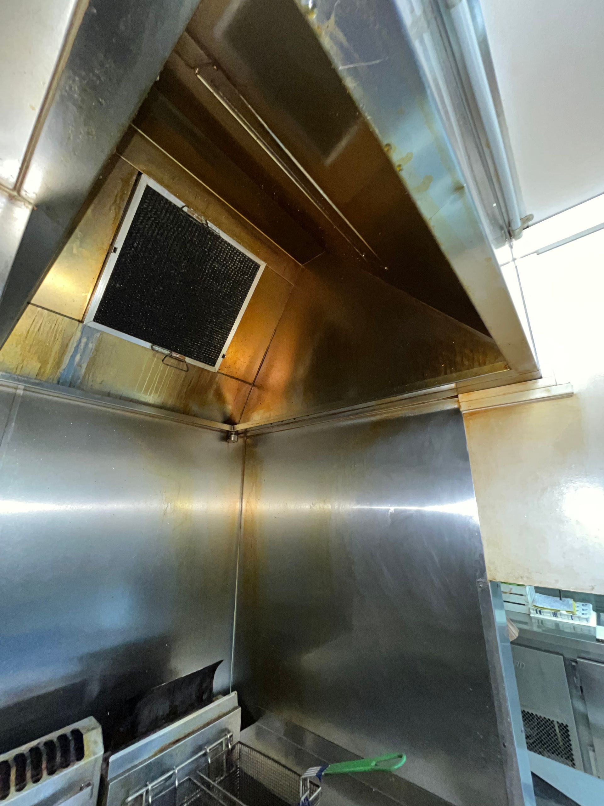 Stainless steel kitchen exhaust hood, interior view. Dark filter, grease stains, and reflective surfaces are visible.  DM & MM Services, Exhaust Cleaning, Townsville, QLD.