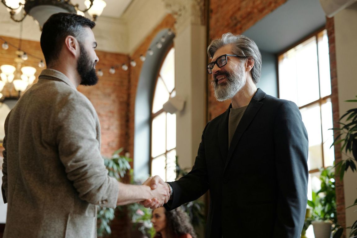Two men shaking hands in a room with brick walls, windows, and plants. One man has a beard and the other has glasses.