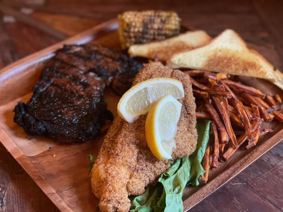 A wooden plate topped with steak , fish , sweet potato fries and corn on the cob.