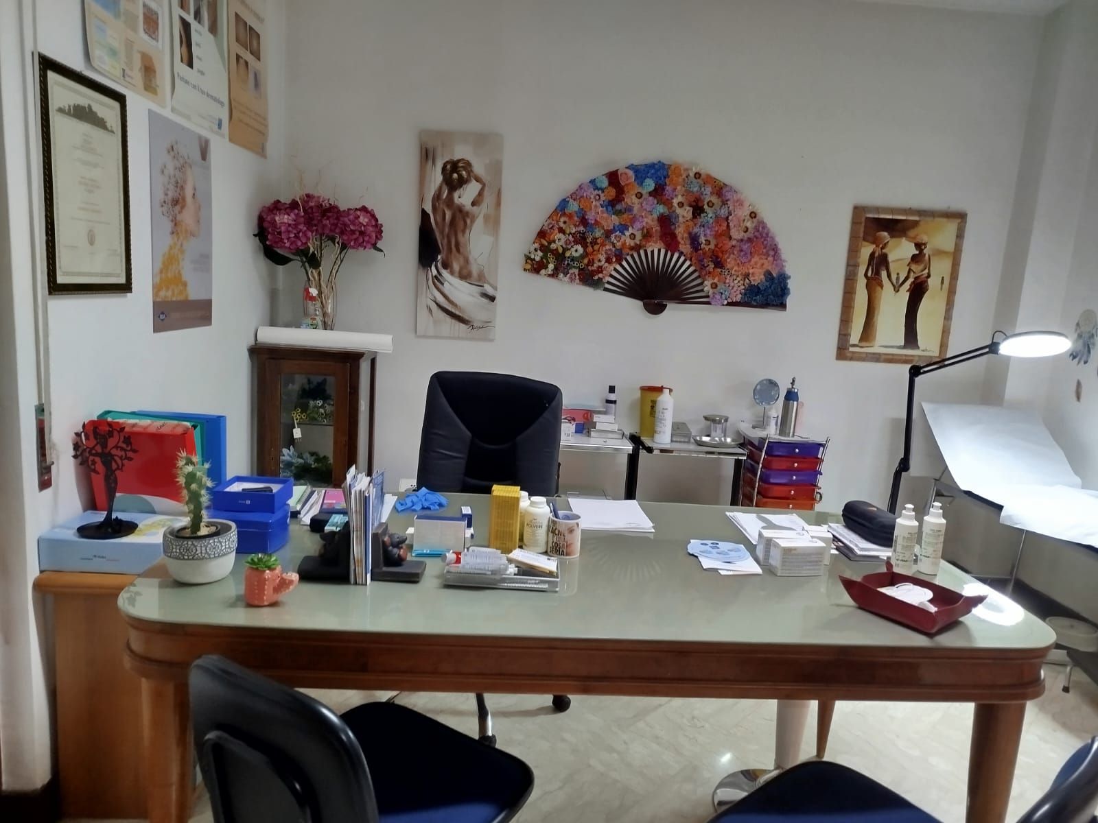 Interno di uno studio medico con scrivania, sedia, lettino per visite, opere d'arte e forniture mediche.
