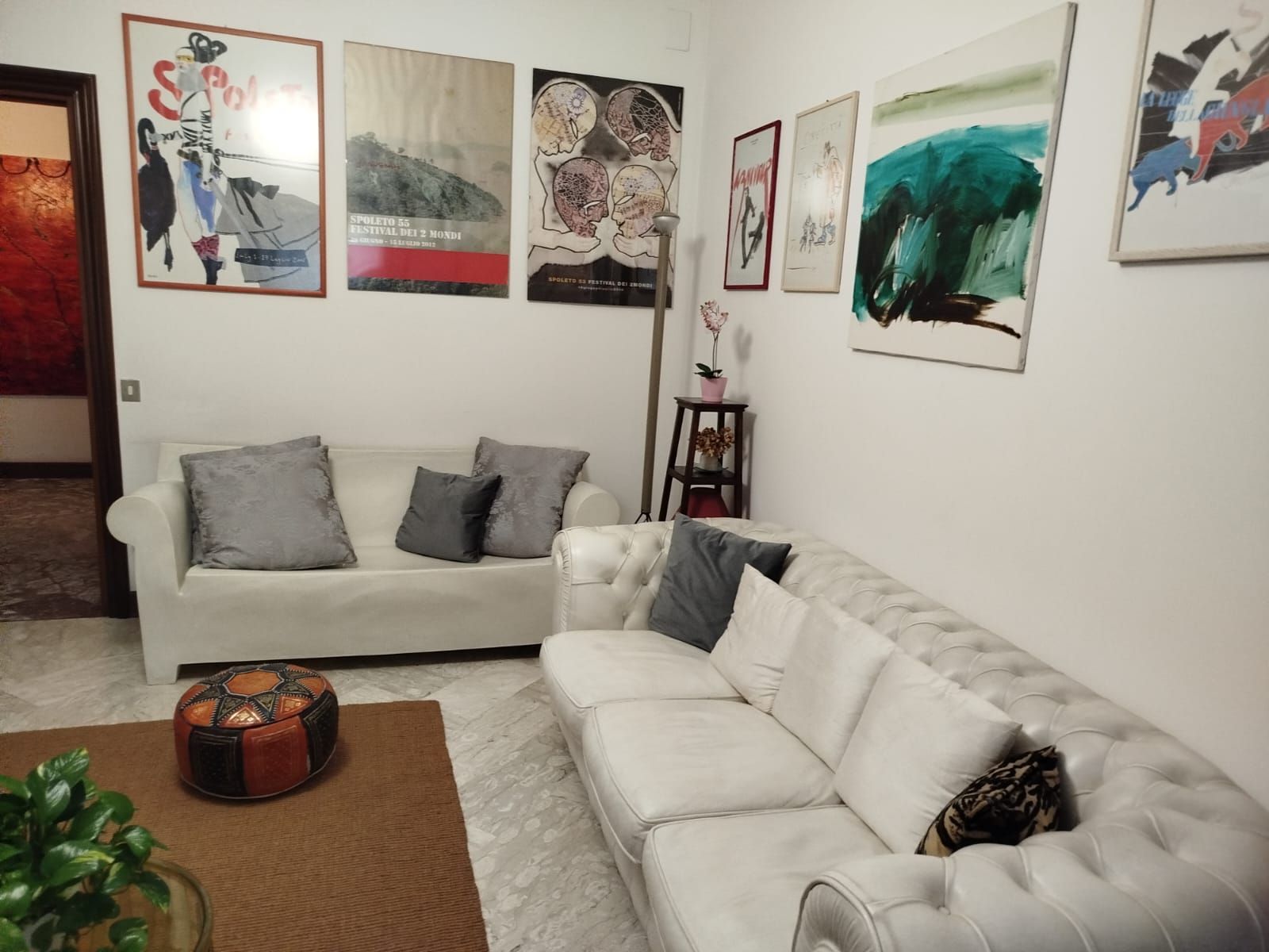 Soggiorno con divani bianchi, opere d'arte alle pareti e un pouf decorato.