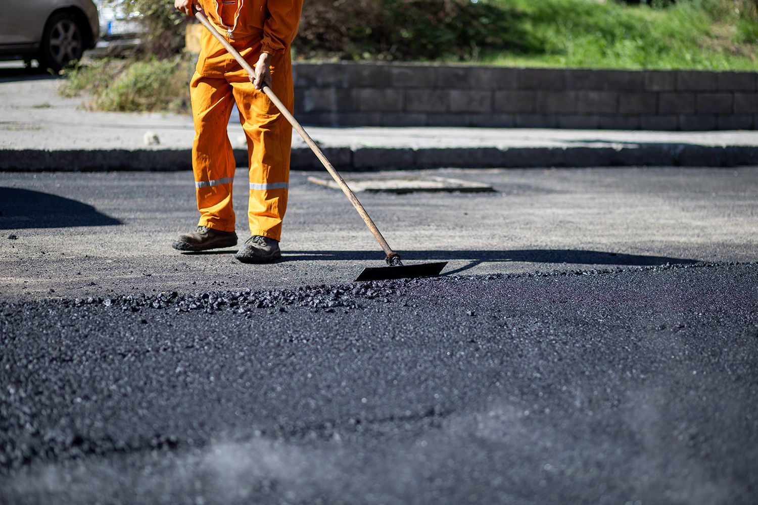 Person in orange jumpsuit smoothing asphalt with a broom on a street.
