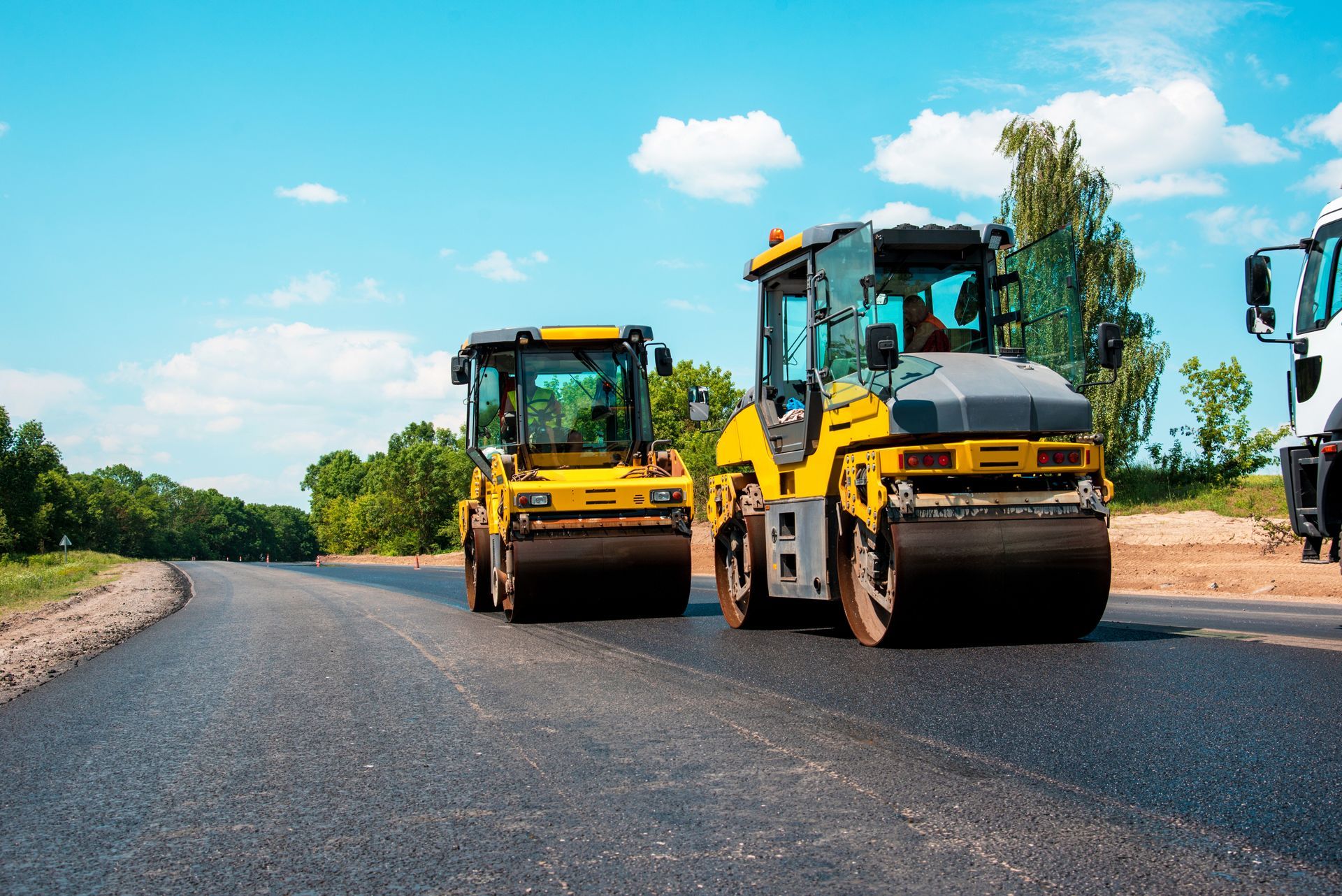 Road rollers compacting fresh asphalt on a sunny road.