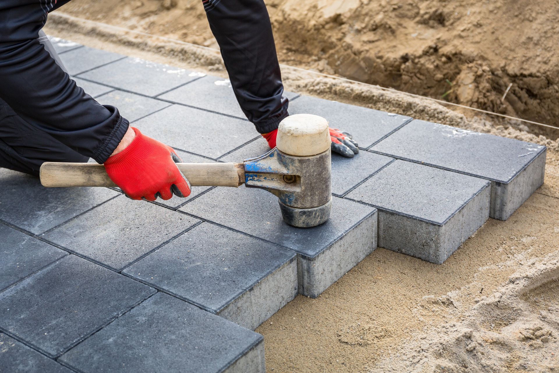 Person in red gloves using a mallet to level dark gray paving stones on a sand base. Person in red gloves using a mallet to level dark gray paving stones on a sand base.