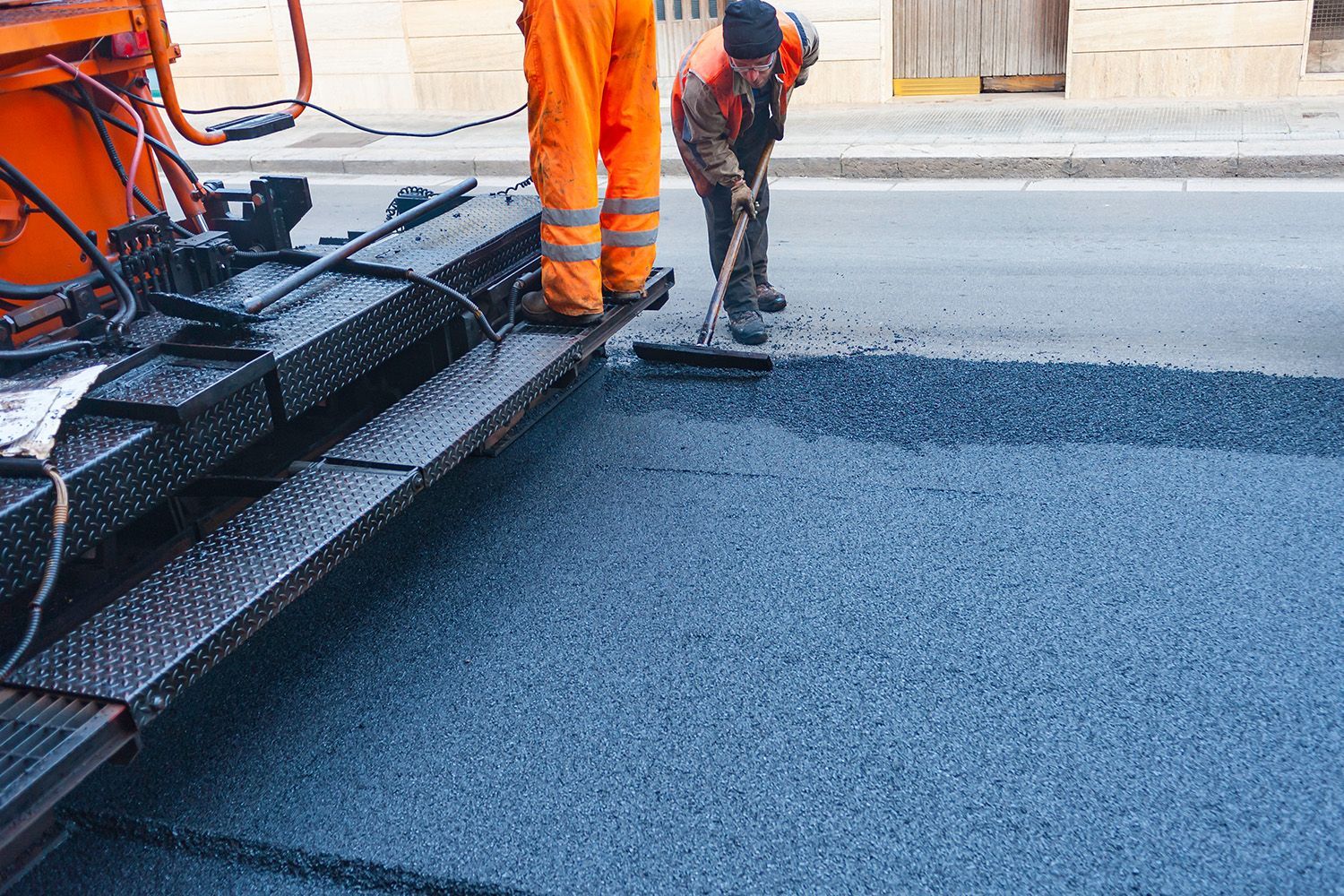 Road workers paving a street with asphalt; one using a broom, the other standing near the machinery.