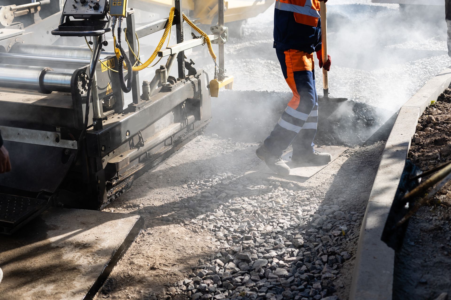 Road construction: Worker in safety gear shovels asphalt near a paving machine laying a sidewalk. Road construction: Worker in safety gear shovels asphalt near a paving machine laying a sidewalk.