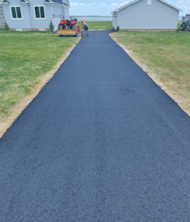 Person in orange workwear uses a long-handled asphalt rake on a newly paved road.