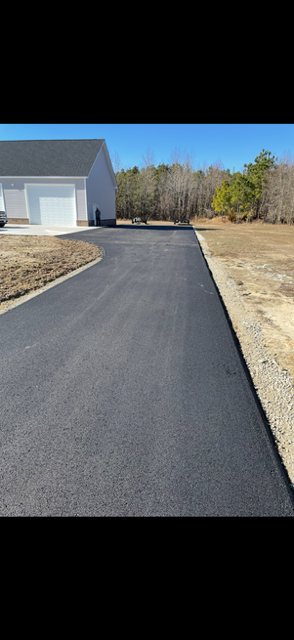 Person applying black sealant to asphalt with a long-handled roller on a sunny day.