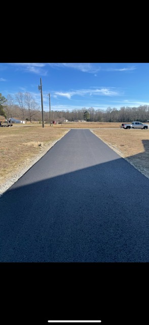 Asphalt paving in progress near a house; worker raking, small roller compacting the fresh asphalt.