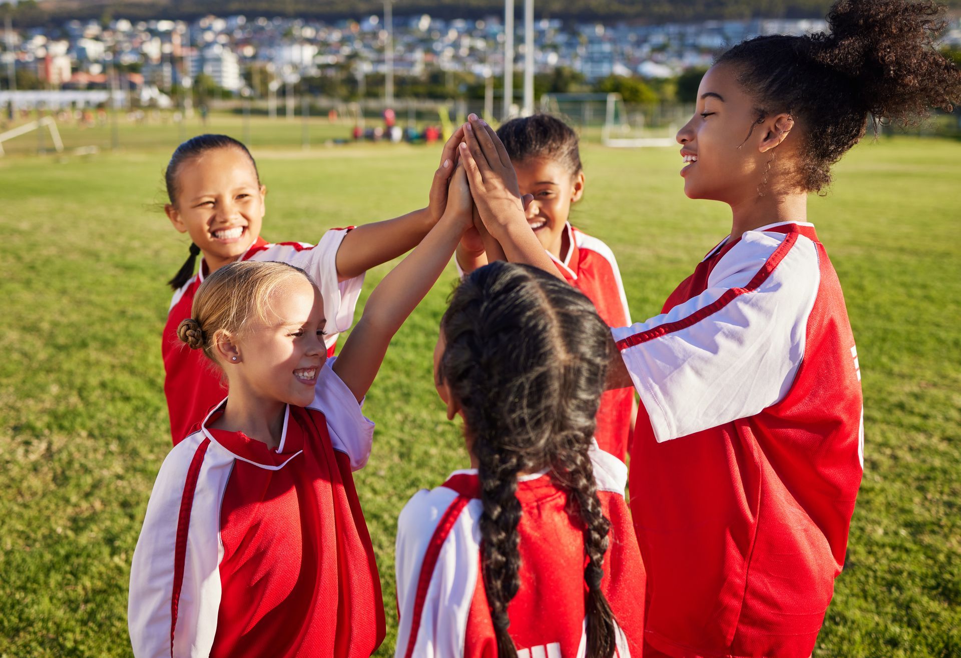 Girls in red and white soccer jerseys high-fiving on a grassy field, smiling.
