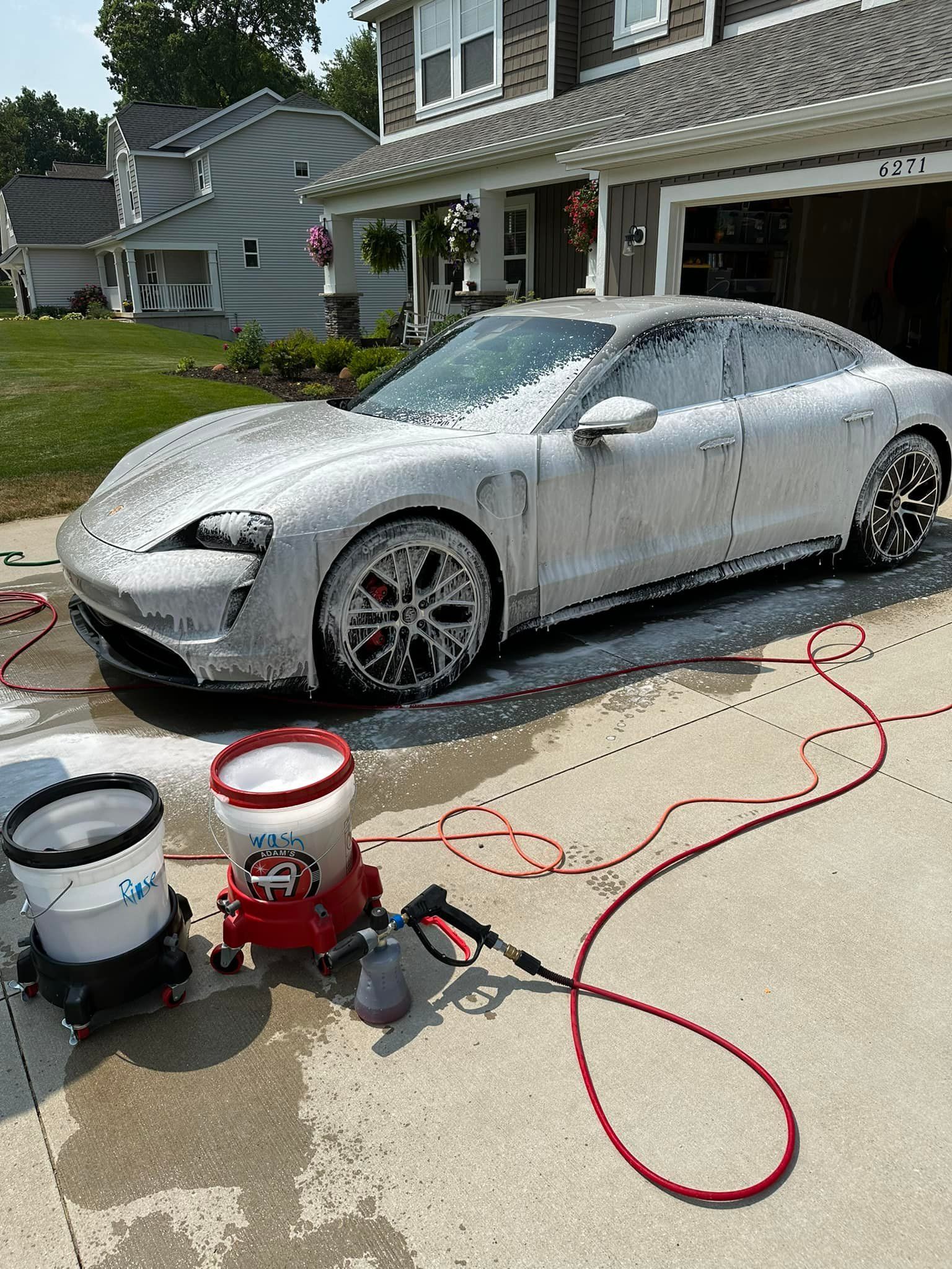 Silver car covered in soap suds in a driveway being washed. Buckets and a soap dispenser are present.
