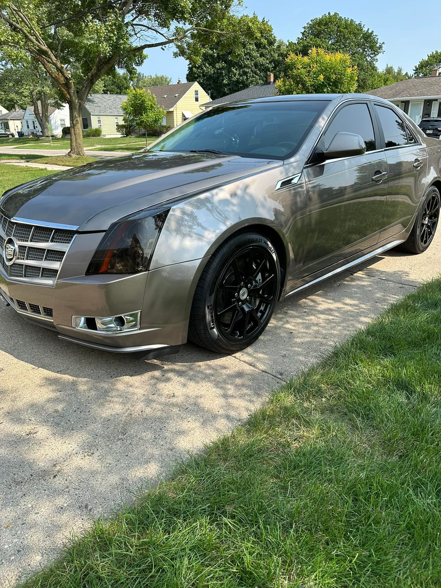 Gray Cadillac CTS parked on a paved driveway with black wheels, tinted windows, and headlight covers, on a sunny day.