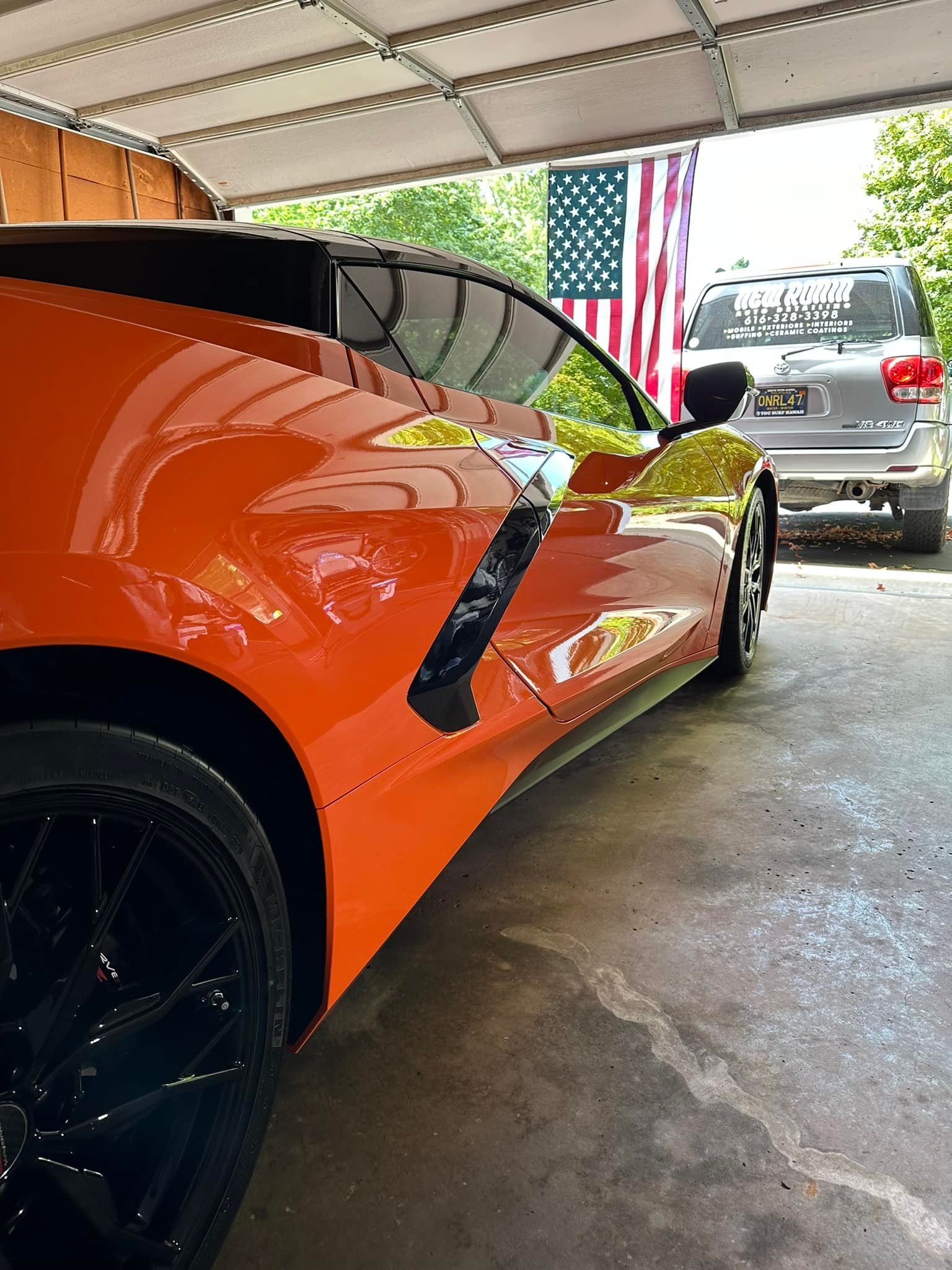 Orange sports car in a garage with a black wheel, an American flag, and a silver SUV.