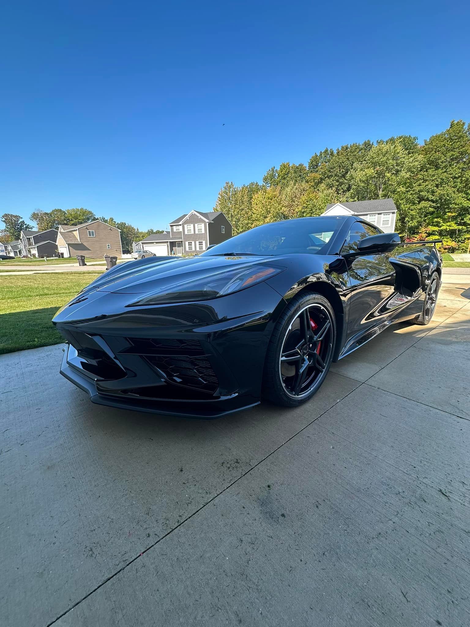 Black sports car parked on a concrete driveway in front of a suburban house on a sunny day.