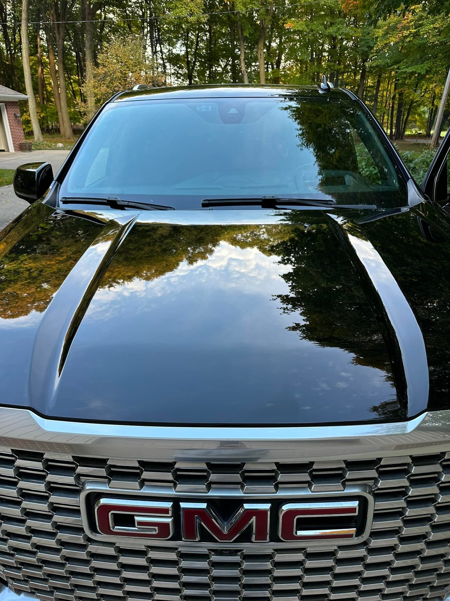 Black GMC SUV, shiny hood reflects trees, chrome grill, red GMC letters.