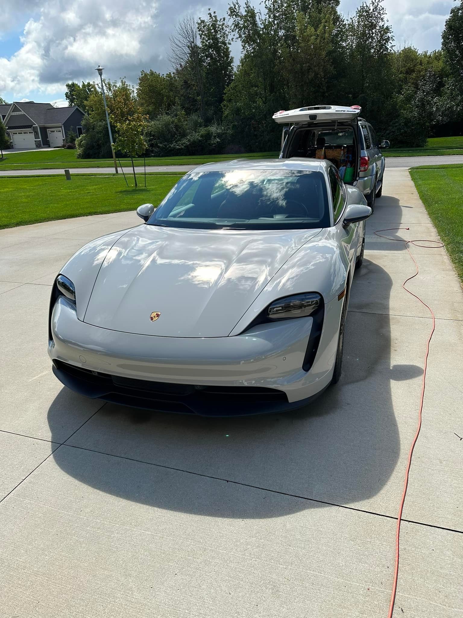 Silver Porsche Taycan parked on a driveway in front of a service cart, sunny day.