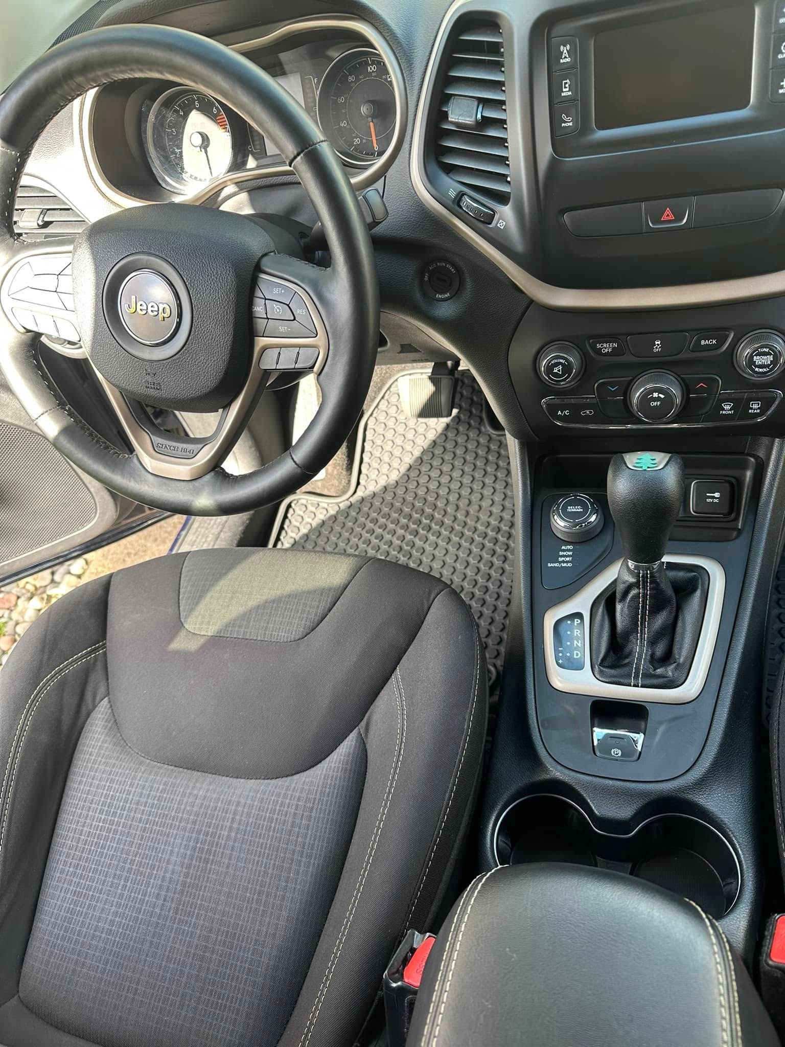 Interior view of a Jeep Cherokee: steering wheel, dashboard, gear shift, and front seat. Black and gray tones.