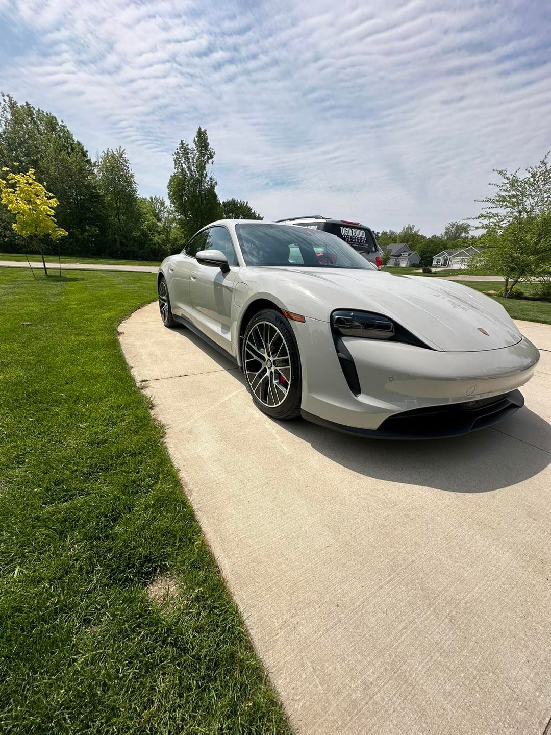 Light gray Porsche Taycan parked on a concrete driveway, surrounded by green grass and trees under a cloudy sky.