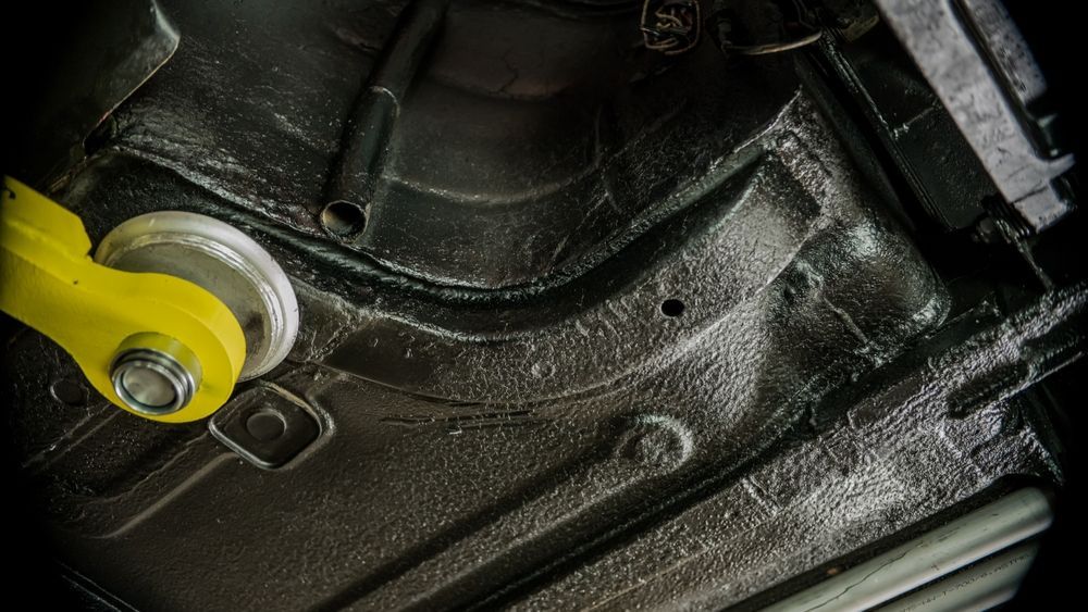 Black undercarriage of a car with protective coating. A yellow lift arm is visible on the left.