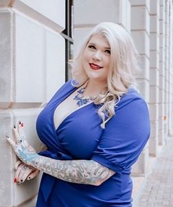 A woman in a blue dress with tattoos on her arms is standing in front of a building.