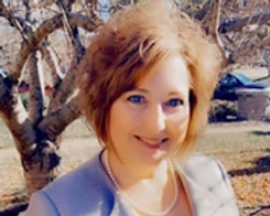 A woman in a suit and pearl necklace is smiling in front of a tree.