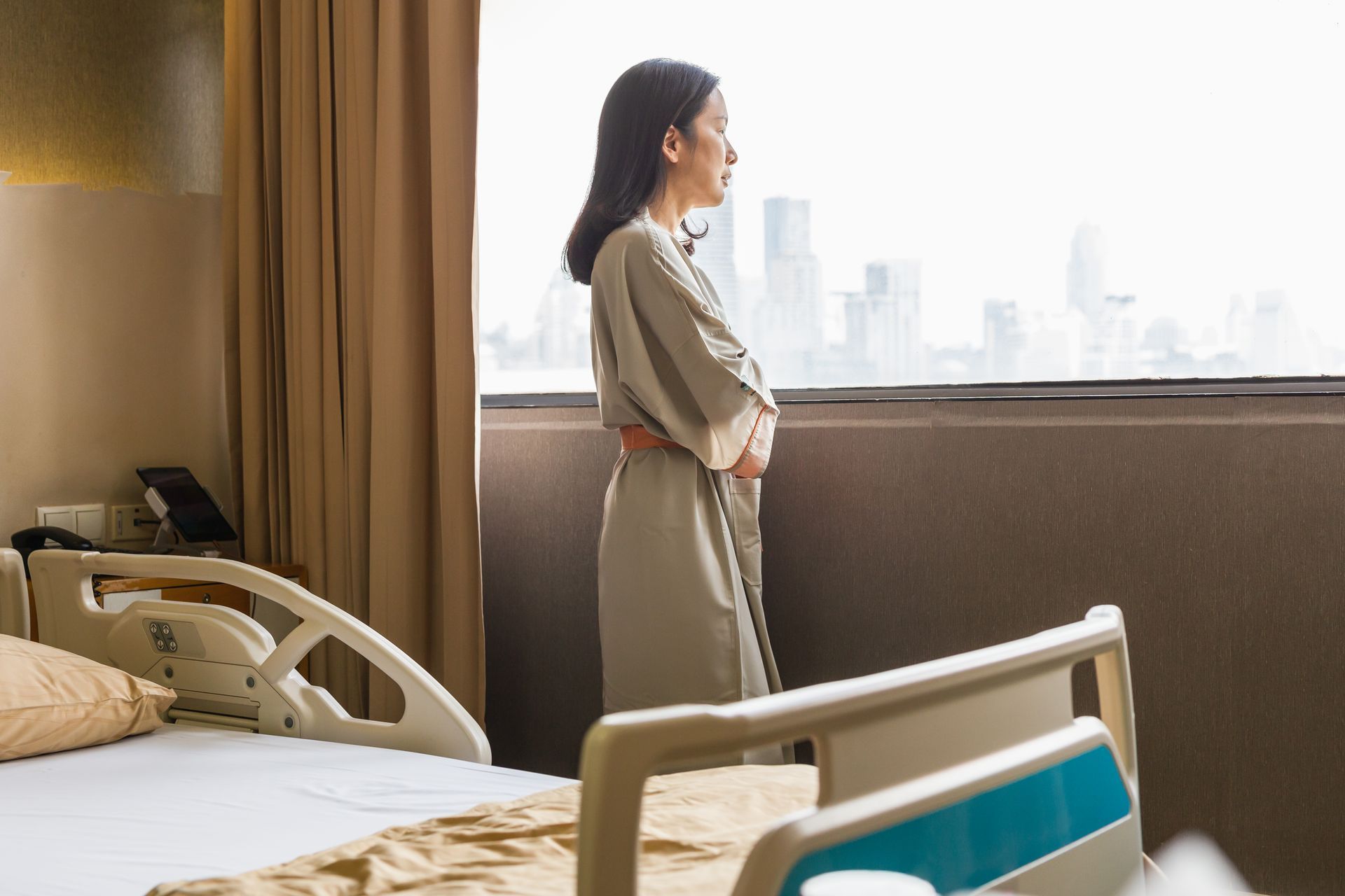 A woman is standing in a hospital room looking out of a window.
