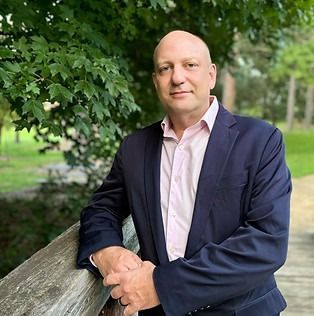 A man in a suit and pink shirt is leaning on a wooden railing.