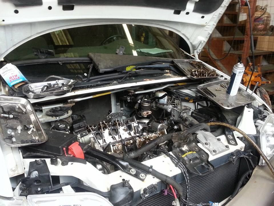The hood of a white car is open, revealing an engine with the cover removed, surrounded by tools in a repair shop. | Auto Service Depot