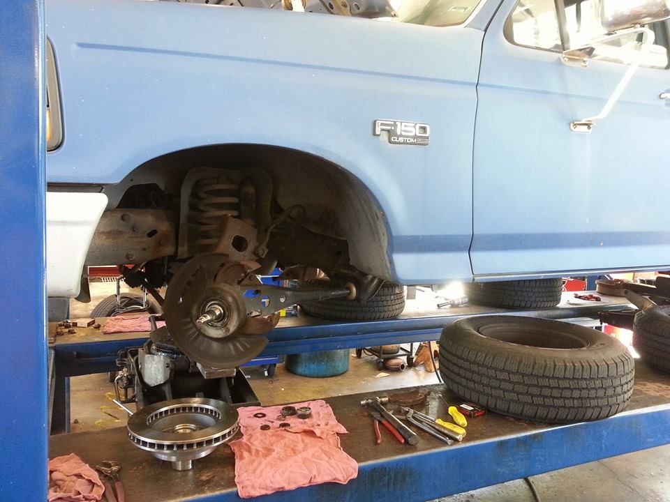 A light blue Ford F-150 truck lifted on a service rack with its front wheel removed for brake maintenance. | Auto Service Depot
