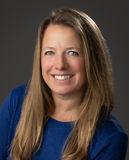 Woman with long blonde hair, smiling, wearing a blue shirt, against a gray background.