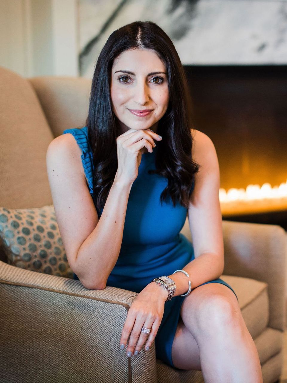 A woman in a blue dress is sitting in a chair in front of a fireplace.