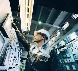 Technician in a hard hat inspecting electrical equipment in an industrial setting.