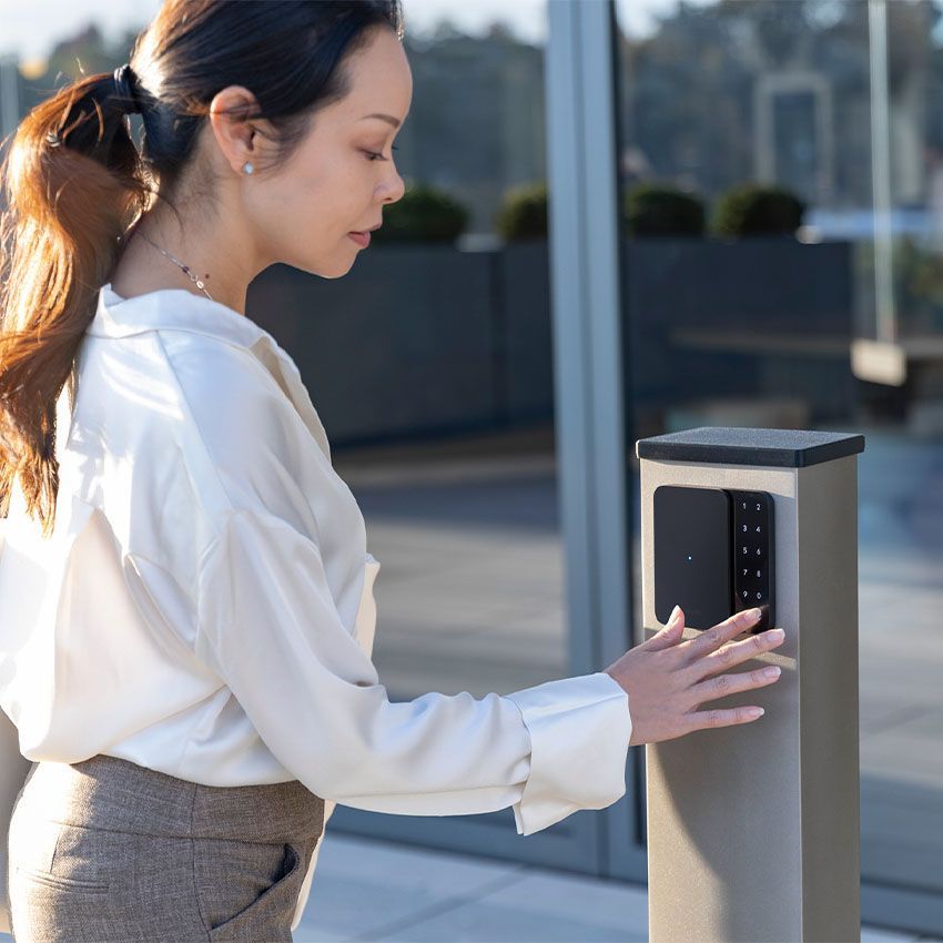 Woman swiping ID card at a time clock on a white wall. She smiles, wearing glasses and a gray sweater.