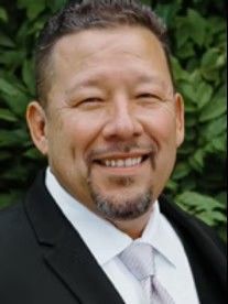 Man in a suit smiling, with a small goatee, against a blurred green background.