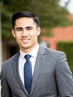 Man in gray suit and navy tie smiles at the camera outdoors.