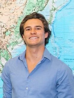 Man smiling, wearing a blue collared shirt, stands in front of a world map.