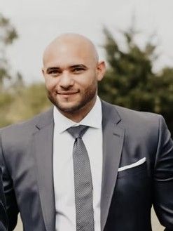 Man in gray suit with a bald head, smiling, in an outdoor setting.
