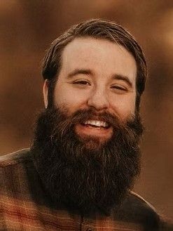 Man with a large, dark beard smiles, wearing a plaid shirt.