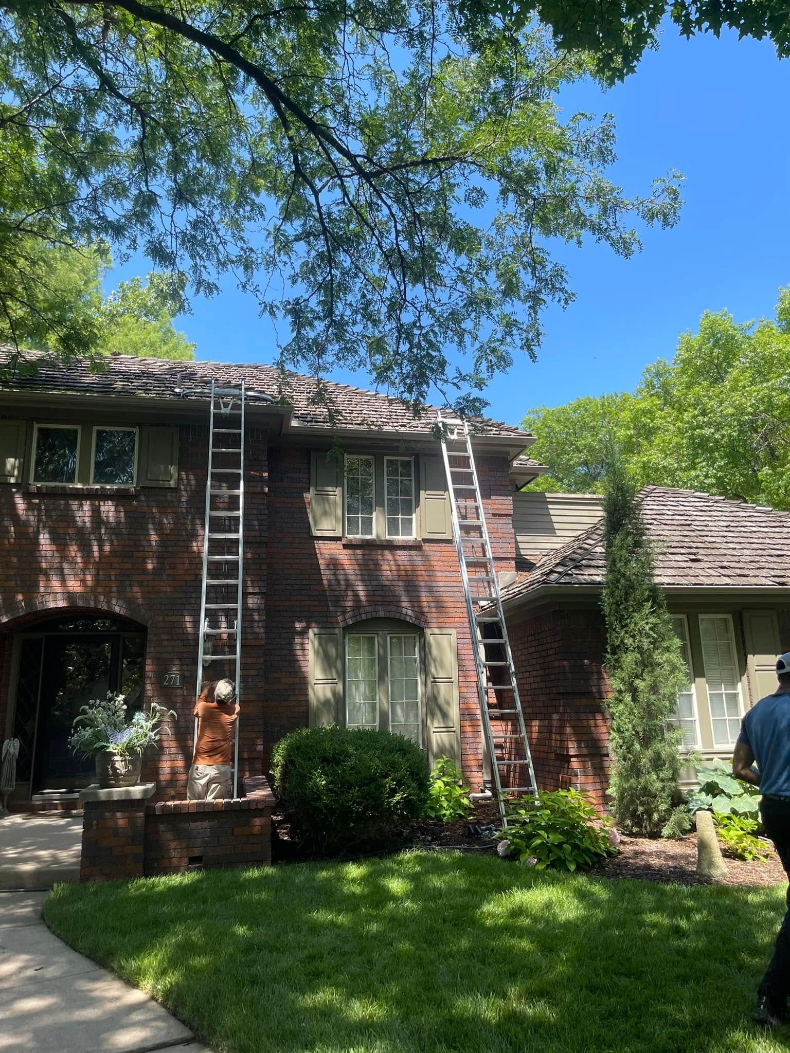 A man is standing in front of a brick house with a ladder on the roof.