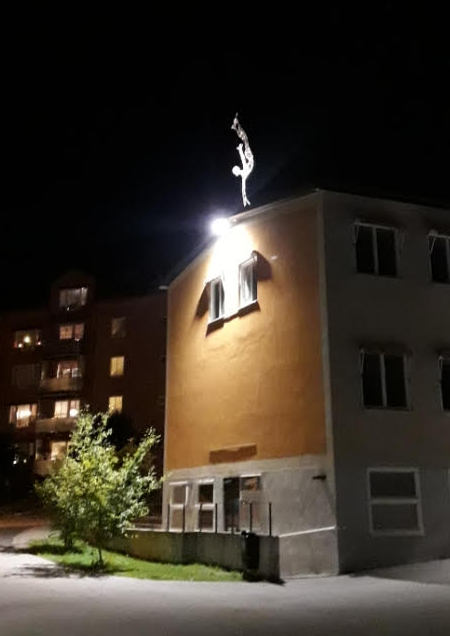 upplyst skulptur i metall i form av en person som står på en hand uppe vid taknocken på ett hyreshus