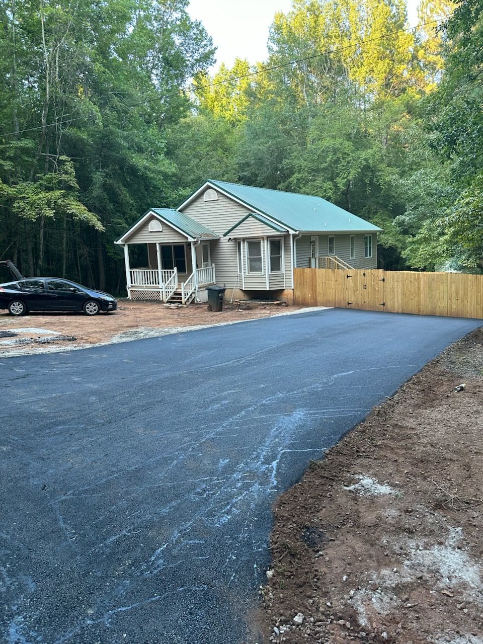 Asphalt driveway leading to a small log cabin with a porch and green roof, surrounded by trees.