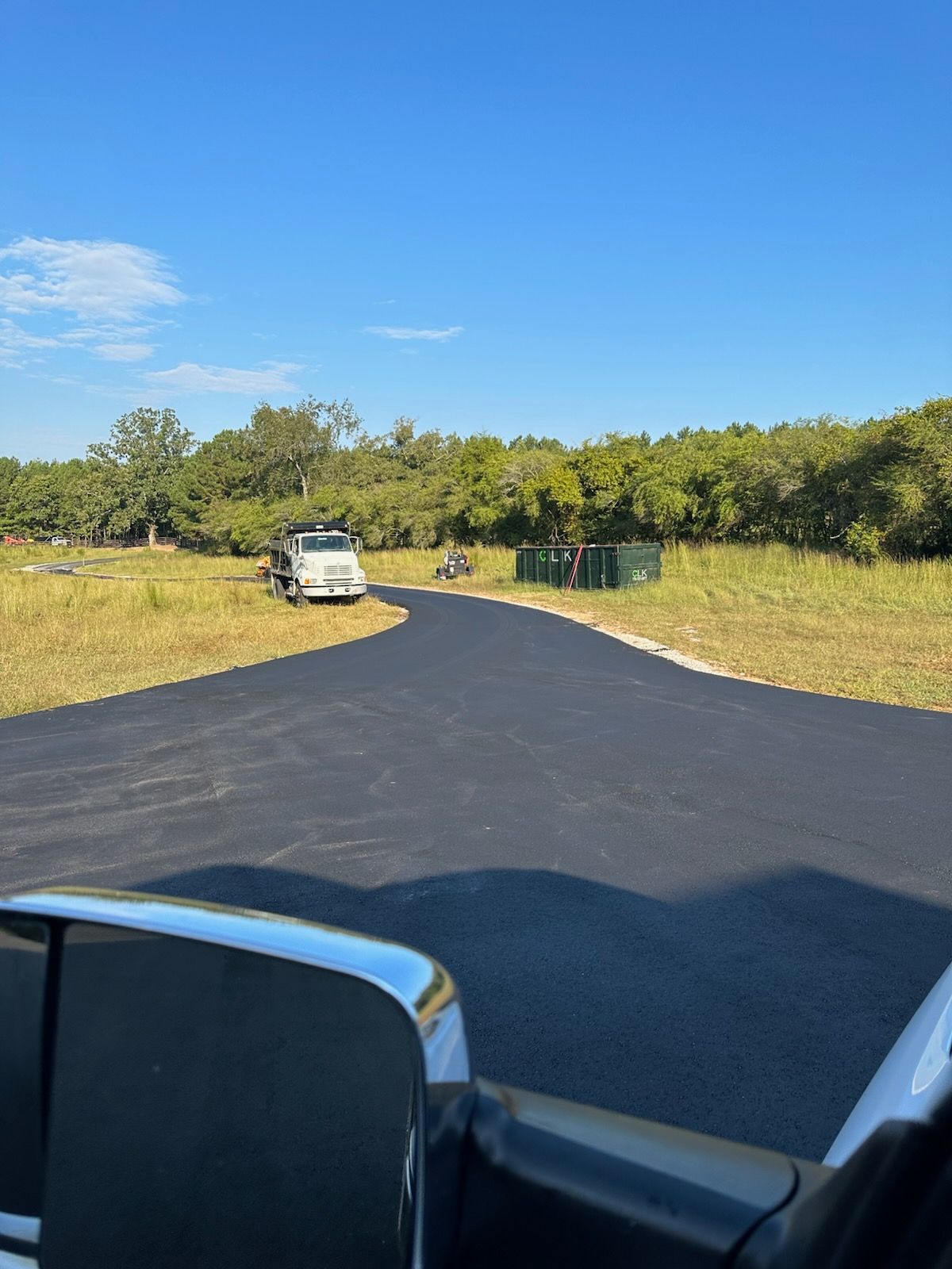 Paved driveway curves into a grassy area under a blue sky, with trees and a truck visible.