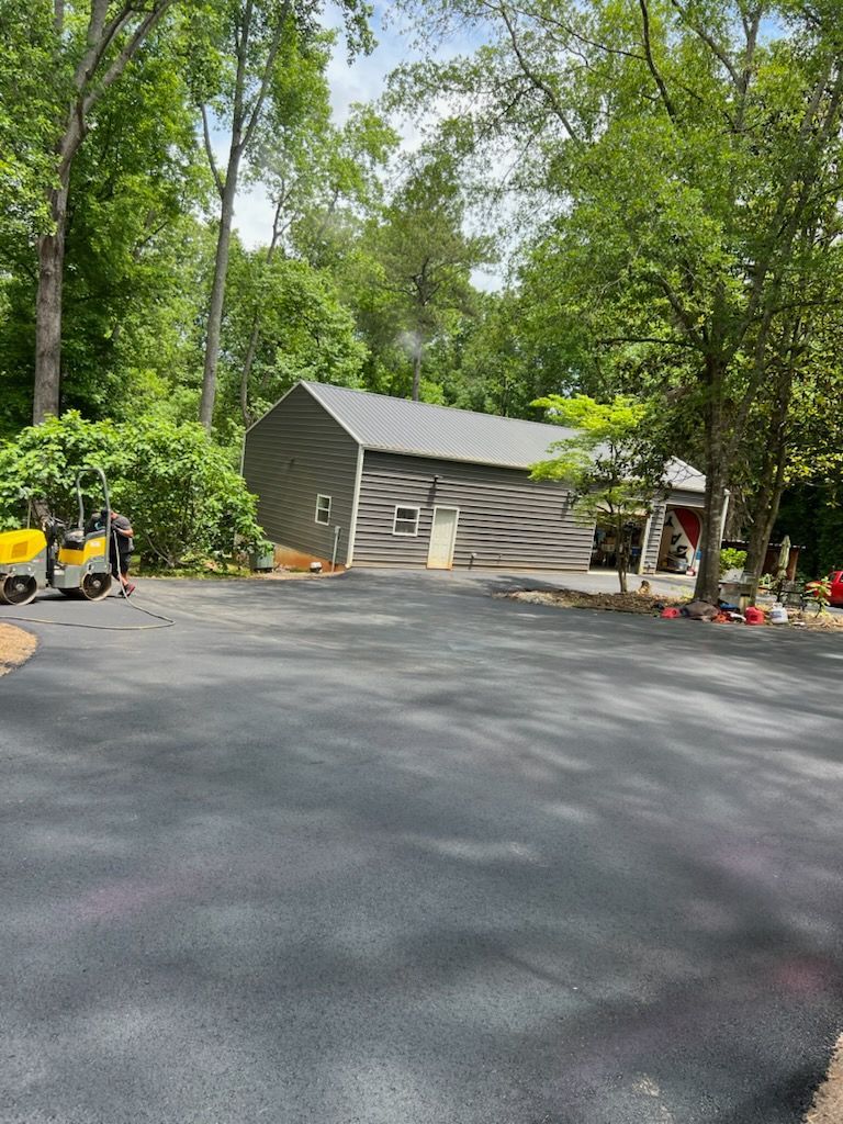 Newly paved asphalt driveway leading to a garage. A person stands near a machine on the left, surrounded by trees.